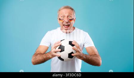Homme caucasien âgé heureux applaudissant avec un ballon de football et maquillage facial, concept sportif Banque D'Images