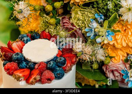 Un gâteau blanc givré garni de baies fraîches est posé sur une table en bois à côté de cupcakes aux baies assortis et de fleurs vives Banque D'Images