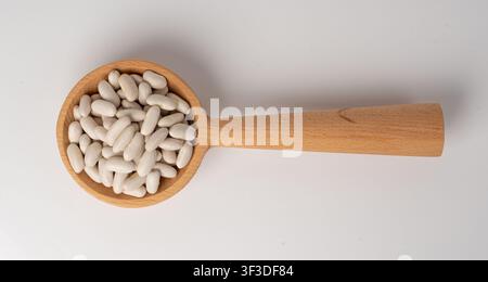 Haricot blanc dans une cuillère en bois isolée, tas de haricots cannellini secs sur fond blanc. Gros plan de haricots blancs crus, graines de lentilles séchées, grains pinto à Banque D'Images
