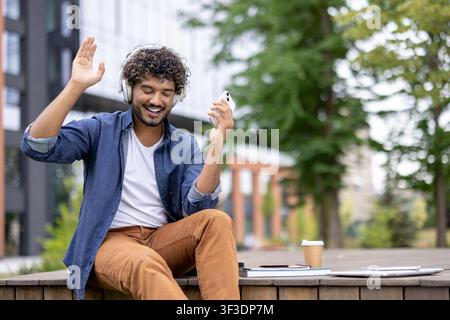 Jeune homme aux cheveux bouclés souriant et faisant un signe de la main, écoutant de la musique via des écouteurs et tenant un smartphone tout en se détendant en plein air, profitant du temps libre et de la technologie Banque D'Images