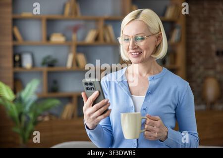 Femme mûre engageant joyeusement avec la technologie, regardant son smartphone tout en se détendant et en savourant une tasse de café chaud dans son bureau moderne à domicile Banque D'Images