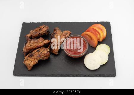 Côtes de porc grillées servies avec une sauce tomate, des tomates tranchées et des oignons sur une planche d'ardoise noire en tant que repas barbecue fraîchement préparé. Banque D'Images