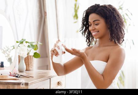 Hydratant la peau. Femme africaine souriante appliquant la lotion corporelle dans la chambre à la maison Banque D'Images