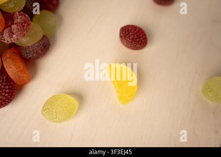 Vue rapprochée de bonbons gélifiés colorés enrobés de sucre et aromatisés aux fruits, éparpillés sur une table en bois clair avec un éclairage doux et chaud et une faible profondeur de champ Banque D'Images