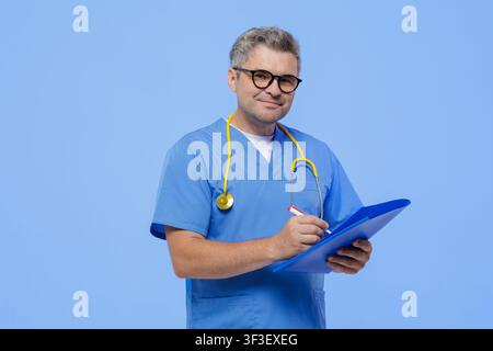 Un médecin professionnel, vêtu de blouses bleues et d'un stéthoscope, écrit des notes sur un tableau à pince en préparation d'un rendez-vous avec un patient dans un établissement médical moderne Banque D'Images