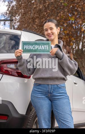 Jeune femme souriante tenant un panneau vert « débutant conducteur » à côté d'un SUV blanc, cadre automnal en extérieur, concept de nouveau conducteur Banque D'Images