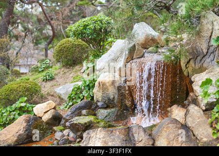 Un jardin japonais serein présentant une petite cascade artificielle dévalant des rochers recouverts de mousse. L'eau coule le long d'une texture brun-rouge Banque D'Images
