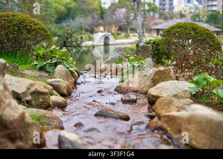 Une scène de jardin japonais sereine mettant en vedette un ruisseau rocheux peu profond s'écoulant vers un étang tranquille au premier plan. De gros rochers lisses bordent le Banque D'Images