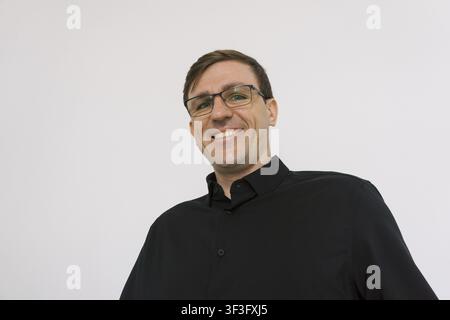 Un homme portant des lunettes et une chemise boutonnée noire sourit avec assurance devant un fond blanc uni Banque D'Images