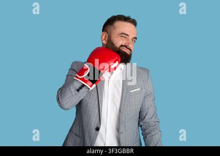 Un jeune homme d'affaires barbu se heurtant au visage avec un gant de boxe rouge, sur fond bleu de studio Banque D'Images