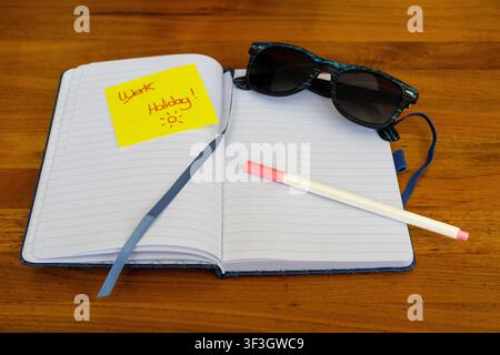 Cahier ouvert avec des lunettes de soleil, un crayon et un post-it jaune portant l'inscription "Vacances", représentant la planification des vacances et la préparation du voyage. Banque D'Images