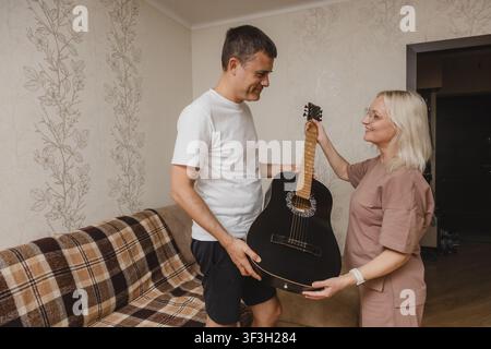 Un homme mûr tend une guitare acoustique noire à une femme mûre souriante dans leur salon à la maison. Banque D'Images
