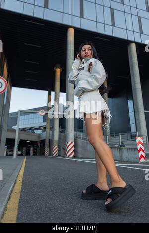 Prise en vue en plein corps d'une jeune femme asiatique confiante dans une tenue en jean blanc tendance et des sandales à plateformes noires, posant sur une rue urbaine devant des corridors modernes Banque D'Images