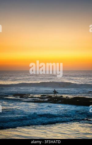 Surfeur marchant sur des rochers de l'océan au coucher du soleil à Imsouane, Maroc Banque D'Images