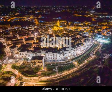 Vue aérienne du centre-ville historique circulaire de Locorotondo la nuit, avec des bâtiments blancs illuminés et le dôme lumineux de la Chiesa Madre di Banque D'Images