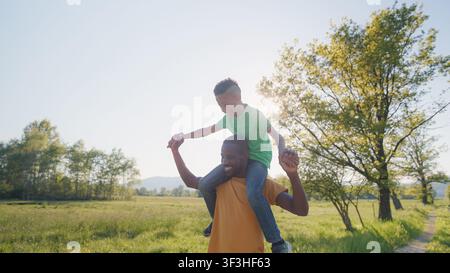 Père porte son fils sur ses épaules alors qu'ils jouent dans un champ pendant l'heure dorée. Ils partagent des rires et de la joie dans la nature. Banque D'Images