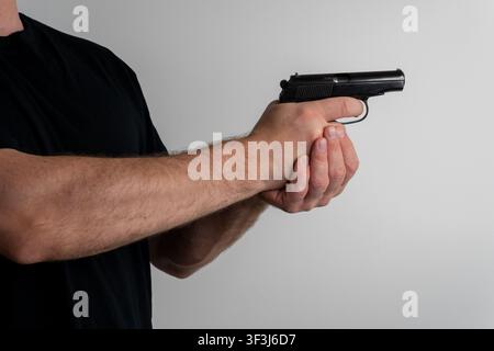Vue recadrée d'un homme en t-shirt noir tenant un pistolet semi-automatique avec une prise à deux mains visant sur fond blanc. Banque D'Images