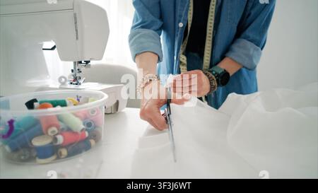 Une personne utilise des ciseaux tranchants pour couper du tissu blanc pour un projet de couture dans un atelier de couture lumineux Banque D'Images