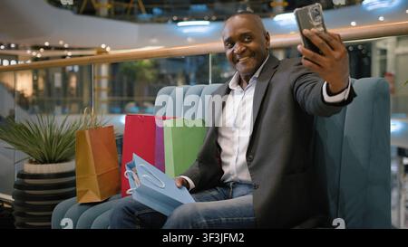 Homme d'affaires mature afro-américain d'âge moyen prenant un selfie avec des sacs de courses à l'aide d'un téléphone portable dans un centre commercial, acheteur ethnique, client, customer Banque D'Images