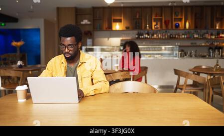Homme afro-américain concentré portant des lunettes travaillant sur un ordinateur portable dans un café et buvant une boisson chaude. Homme assis à une table en bois avec une tasse de café, femme Banque D'Images