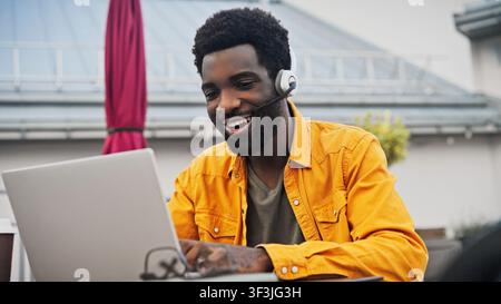 Bel homme afro-américain barbu portant un casque, parlant tout en travaillant sur un ordinateur portable à l'extérieur. Homme communiquant en ligne, assis près d'un espace moderne Banque D'Images