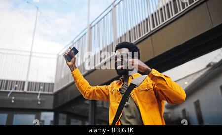 Bel homme afro-américain barbu portant des lunettes tenant un smartphone en plein air, l'air surpris. Homme réagissant émotionnellement à de bonnes nouvelles ou Banque D'Images