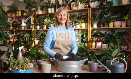 Portrait d'une belle femme mûre attentionnée travaillant dans une fleuristerie et prenant soin des plantes et de la terre. Attrayante femme âgée aux cheveux gris heureuse caucasienne Banque D'Images