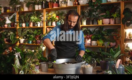 Jeune homme adulte caucasien fleuriste travaillant dans une boutique de fleurs, replantant des fleurs et prenant soin du sol et des plantes. Beau homme concentré aux cheveux longs et barbu Banque D'Images