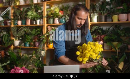 Jeune fleuriste moderne et séduisant travaillant en ligne dans une boutique de fleurs, créant un bouquet de belles fleurs avec un ordinateur portable sur le comptoir. Caucasien attirant Banque D'Images