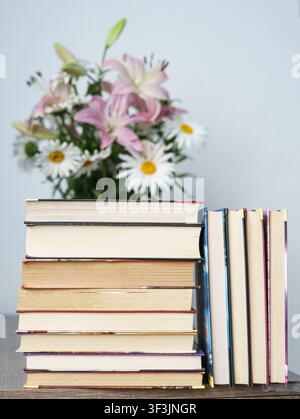Pile de livres colorés sur une table en bois avec un bouquet de lys et de marguerites en fleurs en arrière-plan. Banque D'Images