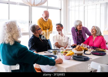 Groupe d'amis seniors divers assis autour d'une table à manger à la maison, discutant et riant avec enthousiasme, célébrant l'amitié et l'âge actif Banque D'Images