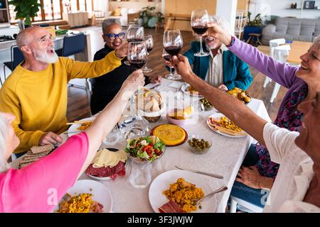 Amis seniors divers et heureux profitant d'un dîner animé à la maison, levant leurs verres de vin ensemble dans un toast joyeux, célébrant la vie et f Banque D'Images