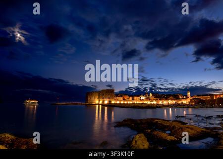 Les anciens remparts de la forteresse et le vieux port de Dubrovnik brillent d'une lumière ambrée chaude à l'heure bleue de la nuit, avec la lune visible à travers les nuages qui se séparent Banque D'Images