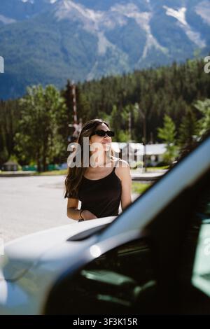 Vue d'une jeune femme portant des lunettes de soleil et un débardeur noir debout à l'extérieur près d'une voiture avec des montagnes et des pins en arrière-plan à Canmore, Banque D'Images