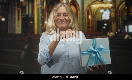 Femme senior blonde pointe le doigt à la boîte cadeau emballée avec la main visible tout en souriant et tenant présent à l'intérieur du bâtiment orné ; contentement chaleureux. Banque D'Images