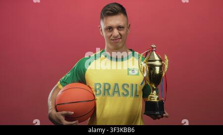 Man in brasil maillot tient basket-ball et trophée d'or avec la main droite tout en grimacant sur fond de studio rouge ; déception. Banque D'Images