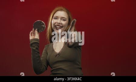Femme tenant un gâteau de riz dans une main tout en tendant l'autre main pour pointer vers l'appareil photo contre un fond studio rouge ; espiègle. Banque D'Images