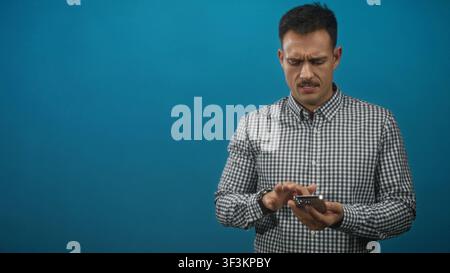 Homme tapant le smartphone avec l'index tout en tenant le téléphone dans les mains sur fond bleu dans le studio ; concentration de frustration. Banque D'Images