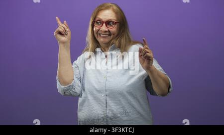 Sénior femme pointe ses deux index vers le haut et sourit largement, portant des lunettes rouges et une chemise à carreaux clairs en studio ; célébration de joie. Banque D'Images