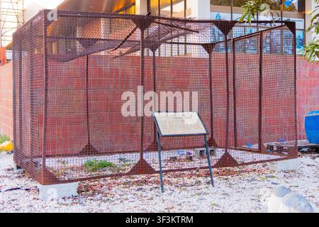 Lusaka, Zambie - 01 juillet 2026 : Vue d'une exposition historique de cage à prisonniers en grillage métallique rouillé, posée sur un sol de gravier devant un rouge Br Banque D'Images