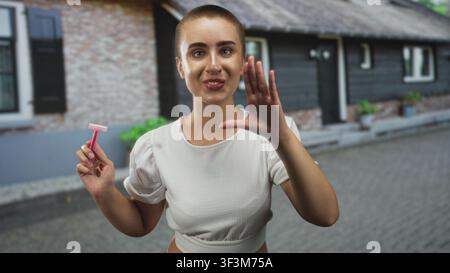 Jeune femme souriante aux cheveux courts tenant un rasoir rose et levant sa main droite, paume vers l'avant, dans la rue ; soins personnels ludiques. Banque D'Images