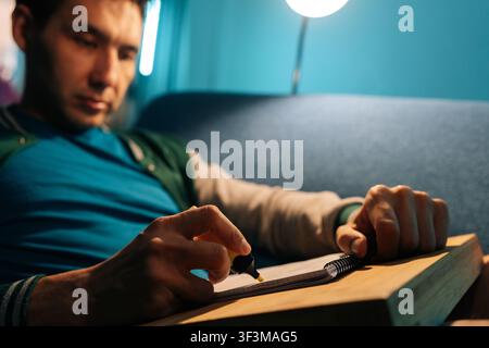 Jeune homme se concentrant sur ses études à la maison pendant la nuit, soulignant des informations importantes dans un cahier à spirale, éclairé par une lampe de bureau Banque D'Images