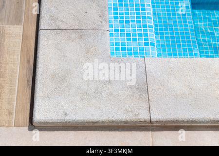 Vue détaillée de la transition entre une terrasse en bois et un rebord de piscine en granit ainsi que des marches carrelées en bleu menant à une eau claire Banque D'Images