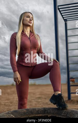 jeune femme blonde athlétique déterminée en tenue de sport mauve assortie, debout avec son pied sur un pneu d'entraînement dans un parc de fitness en plein air sous un ciel nuageux. Banque D'Images