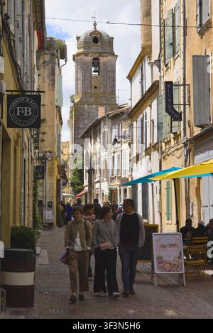 Les piétons marchent dans une rue historique en direction d’un clocher d’église traditionnel au charmant centre d’Arles, en France. Banque D'Images