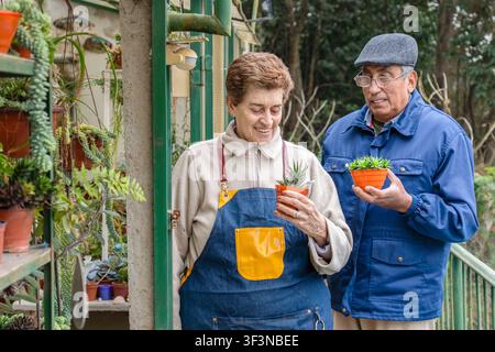 Une femme mûre vêtue d'un tablier en jean et un homme tiennent chacun un petit pot orange contenant des succulentes haworthia devant leur serre, entourés d'un jardin luxuriant Banque D'Images