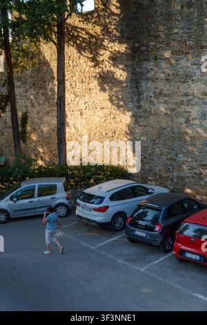 Florence, Toscane, Italie. 28 mai 2026. - Une femme marche à travers un parking goudronné sous de grands arbres et un ancien mur de pierre pendant la dorée Banque D'Images