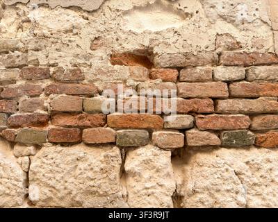 Un gros plan d'un mur en plâtre vieilli révèle une tache de brique rouge exposée parmi des pierres naturelles rugueuses, usées par des décennies de temps. Banque D'Images