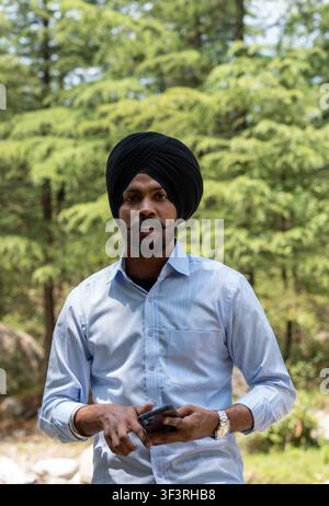 Prise verticale d'un jeune homme sikh portant un turban noir et une chemise bleue, tenant un smartphone dans ses mains tout en regardant directement l'appareil photo. Banque D'Images