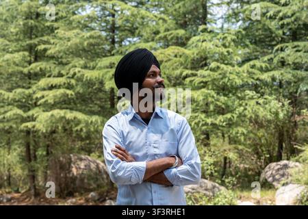 Un jeune Sikh souriant portant un turban noir, debout à l'extérieur les bras croisés, regardant au loin devant un fond d'arbres verts à Himachal. Banque D'Images